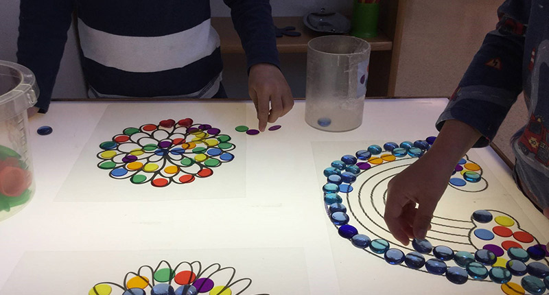 Vitt bord med ritade blommor och regnbåge. Barnhänder fyller figurerna med färgade runda stenar.