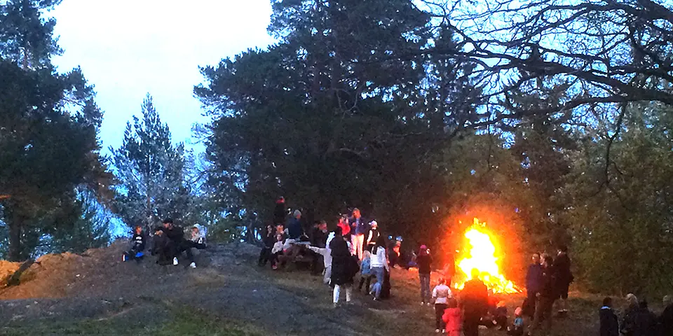 Människor står och tittar på en valborgsmässoeld. Det är skymning ute.