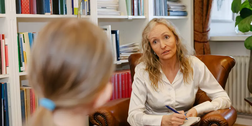 En kvinna sitter i en fåtölj och lyssnar på en annan person som vi bara ser huvudet bakifrån på.