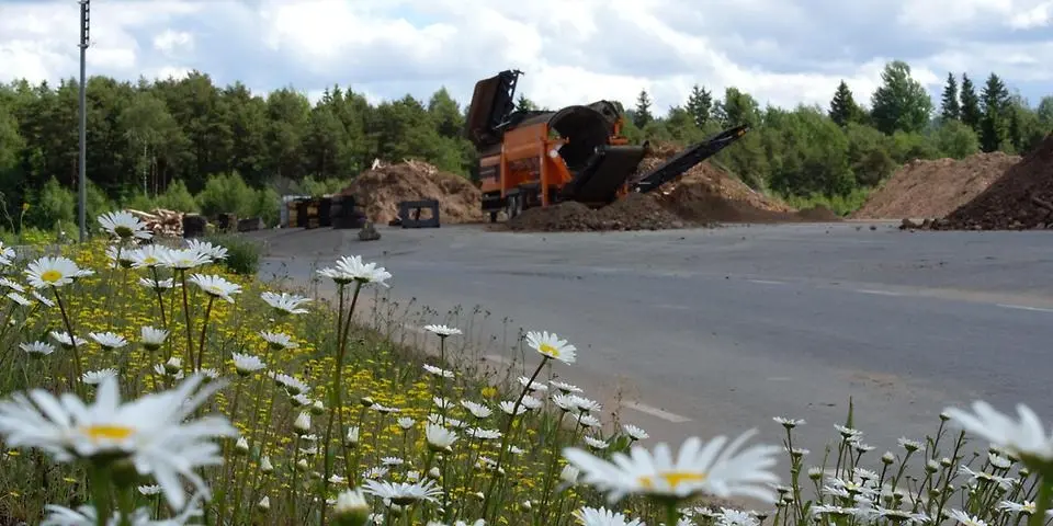 I förgrunden ser du en blomsteräng. I bakgrunden är det en grävmaskin som gräver i en jordhög.