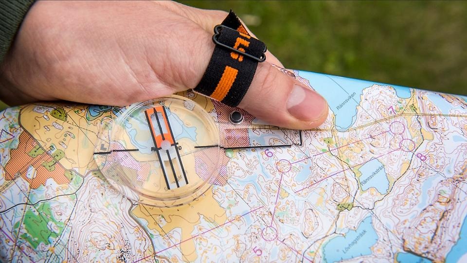 En hand håller i en orienteringskarta med en tumkompass, redo för orientering i skogsterräng.