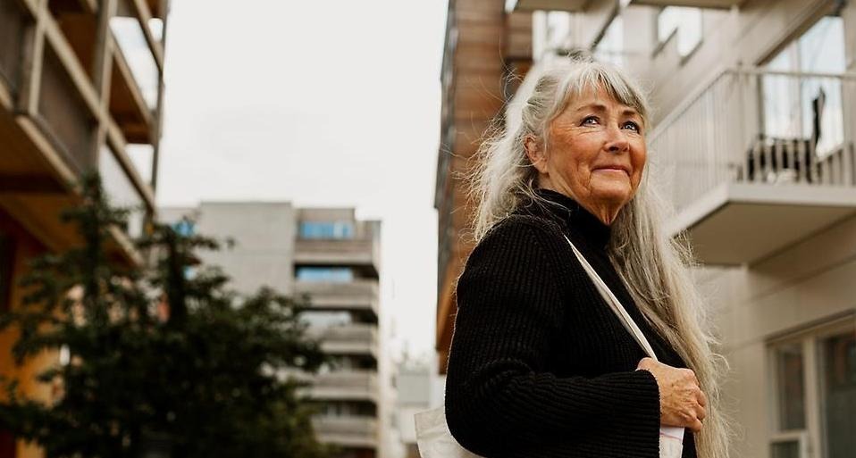 Leende kvinna, långt grått hår, svart jacka, vit axelväska, i bakgrunden lägenhetshus. 