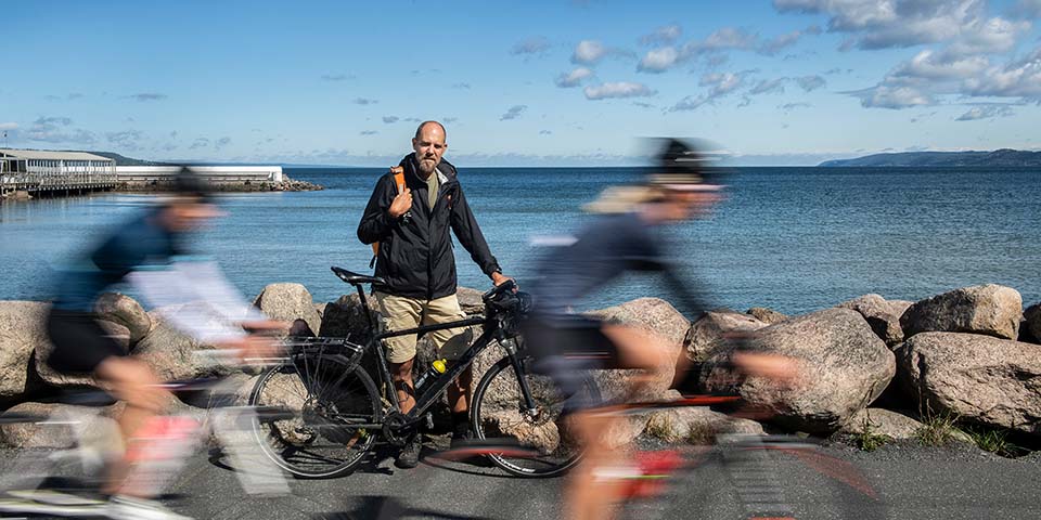 Person står med cykel vid Vättern, två cyklister kör snabbt förbi.