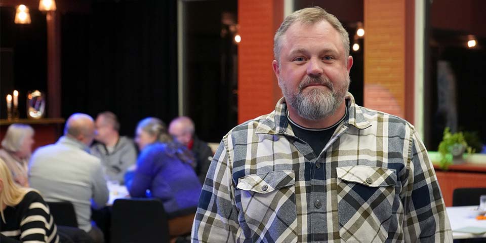 Thomas Olsson tittar in i kameran och ler. I bakgrunden sitter människor och diskuterar runt ett bord.
