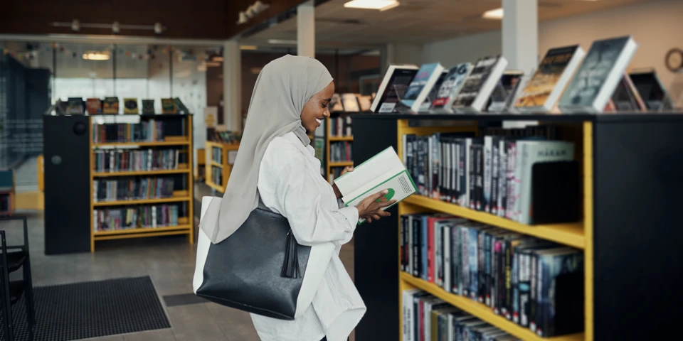 Kvinna med slöja och hörlurar läser bok på bibliotek. 