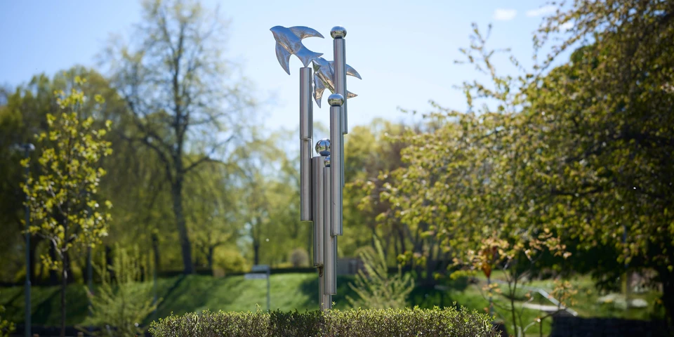 Aluminiumskulpturen omringad av gröna träd och buskar.