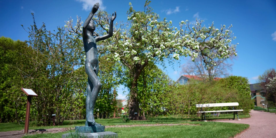 Skulptur i brons som lyfter händer mot himlen, omgiven av blommande träd i en park.