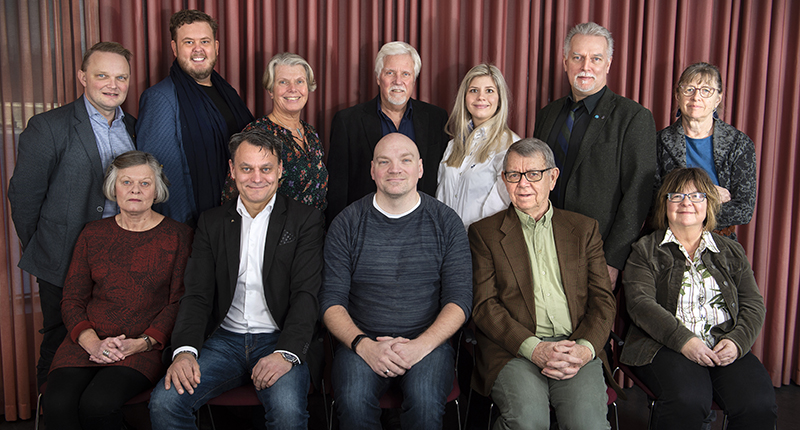 Gruppbild på män och kvinnor framför rostrött draperi.