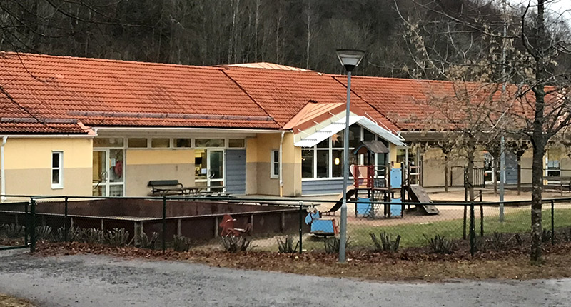 Enplansbyggnad, en förskola med gul fasad och rött taktegel. Lekplats framför med rutschkana och inhängnad rink.
