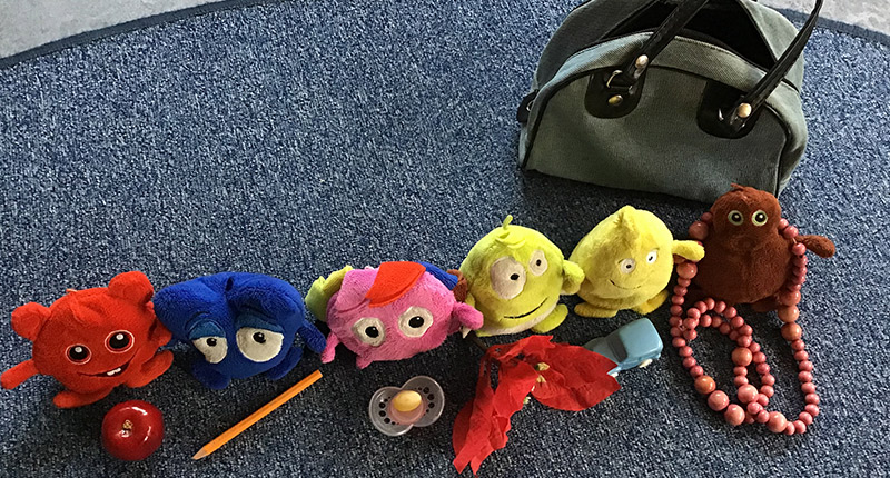Små färgglada figurer uppställda på en blå matta. I bakgrunden en grå bag.