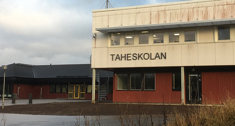 Stor tvåvåningsbyggnad med röd undervåning och vit övervåning. På den vita fasaden står Taheskolan med svarta bokstäver. Till vänster i bild, strax bakom syns en röd enplansbyggnad med svart tak.