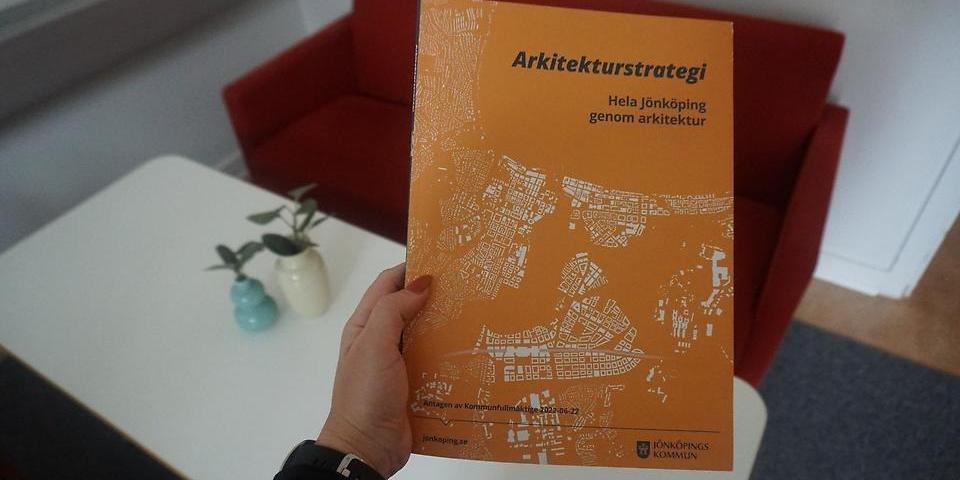 En hand håller i arkitekturstrategin i katalogformat. I bakgrunden syns ett bord med växter och en soffa.