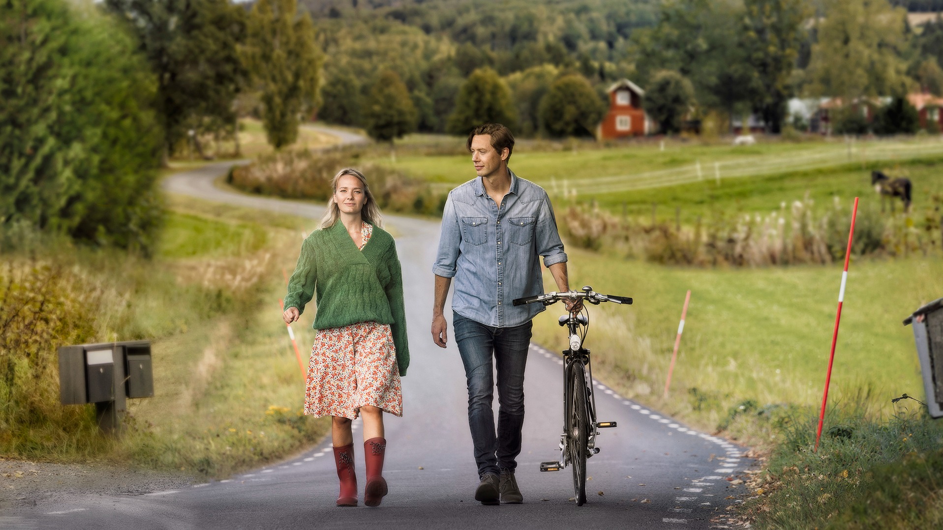 Bilden föreställer de två karaktärerna Lena och Daniel som går på en landsväg i ett grönskande landskap. Daniel leder en cykel.