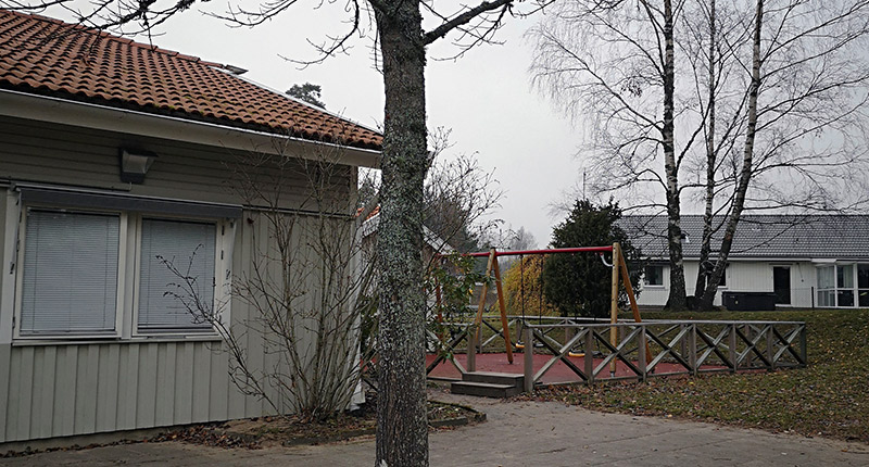 Till vänster syns hörnet på en vit enplansbyggnad. Strax till höger ligger en lekplats med gungställning.