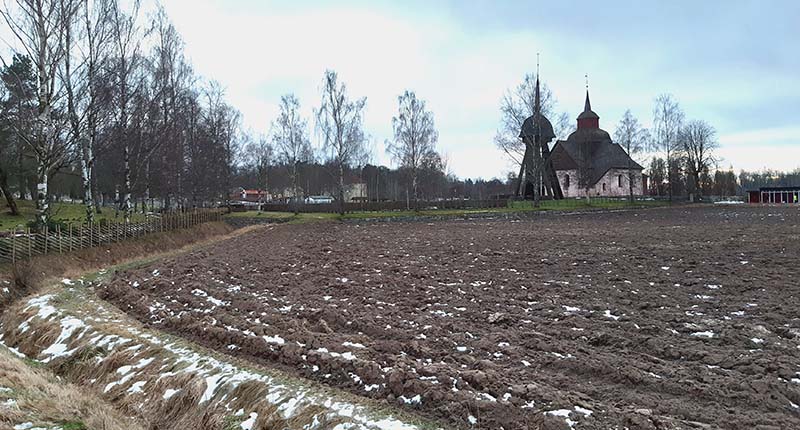 En åker med snöfläckar, i bakgrunden en vit kyrka med klockstapel. 