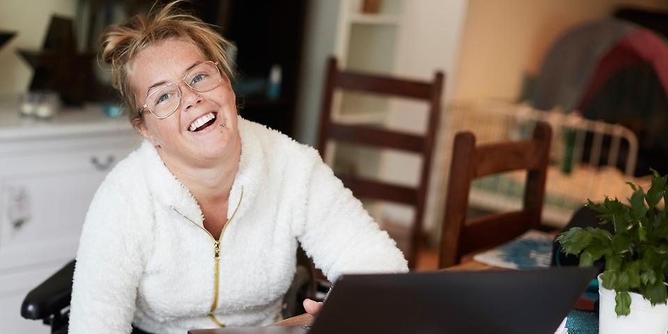 Leende kvinna, ljust hår i toft, glasögon, vit stickad tröja med dragkedja, laptop på bord.