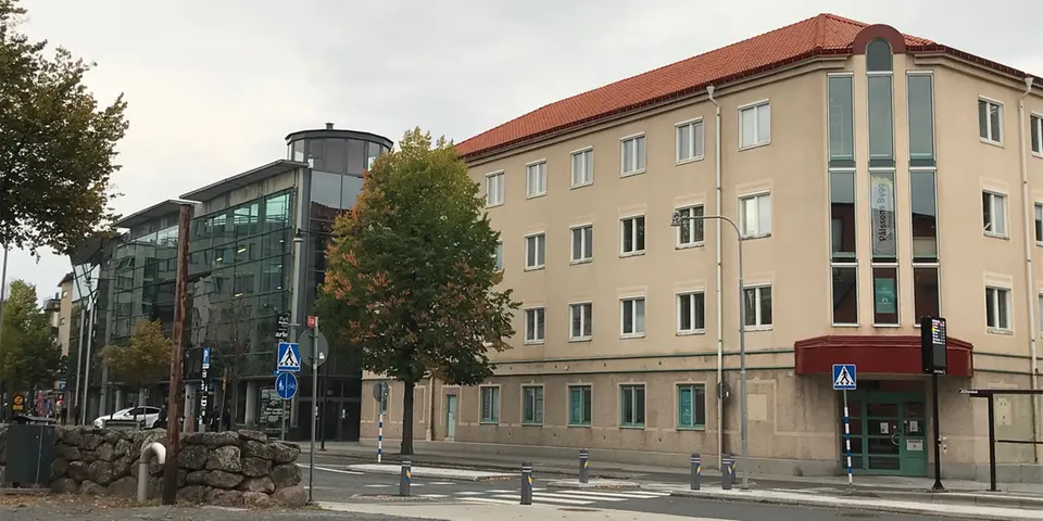 Gult putsat hus med fyra våningar, rödentré och rött tak. Gata med övergångsställe och busshållplats utanför. Parkeringshus i mörkt glas jämte.