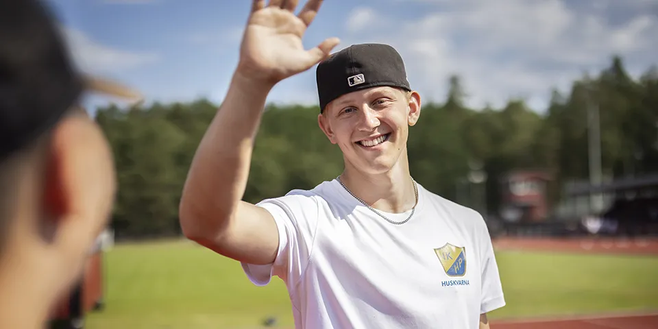 En ung kille med bakåtvänd keps ler emot kameran och håller upp sin hand för att göra en high five. I bakgrunden syns en idrottsplats med grönt gräs och löparbana.