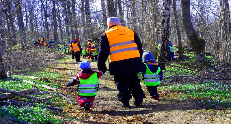 Förskolegrupp på promenad i skogen. Förskolelärare håller två barn i handen.