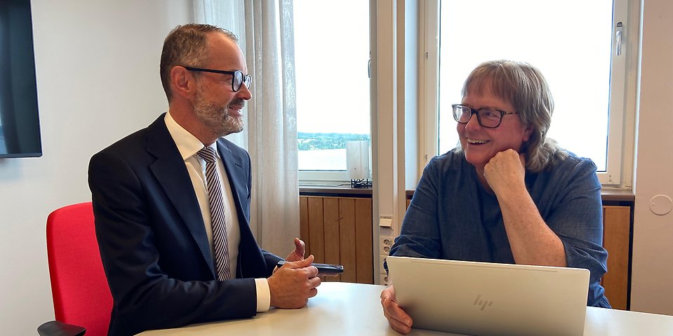 En man med glasögon och kort skägg som har mörk kavaj,
vitskjorta och slips sitter och pratar med en kvinna med lugg och glasögon som
har en mörk tröja. Bakom finns fönster och på bordet framför dem står en bärbar
dator.