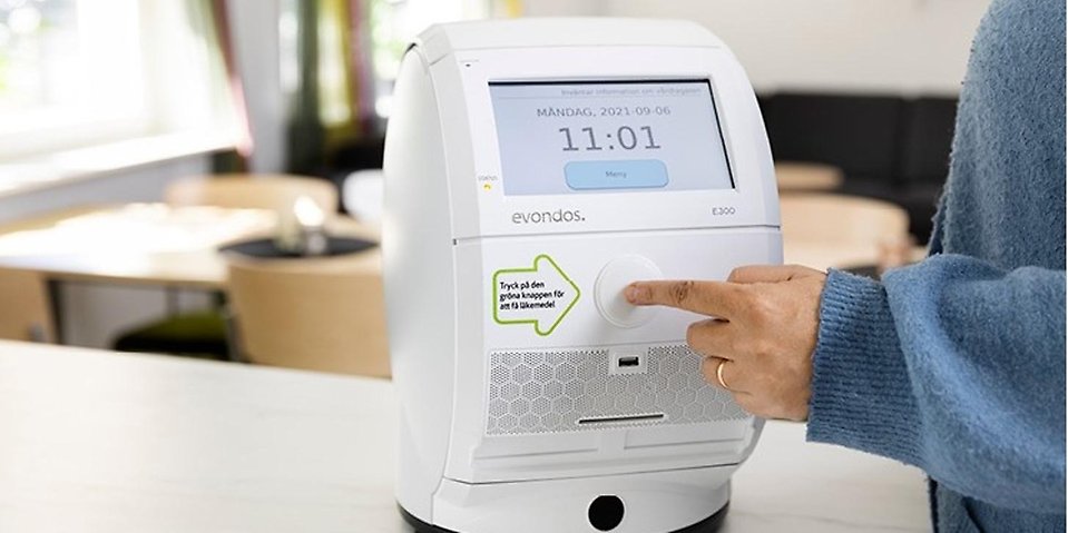Arm med blå tröja, ett pekfinger som trycker på en knapp på en vit maskin med display, i bakgrunden syns ett bord.